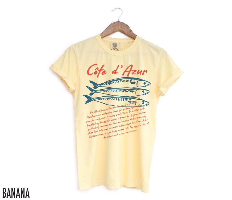 May include: A light yellow t-shirt with the text "C&ocirc;te d'Azur" in red and three blue fish illustrations. Below the text is a paragraph of red text. The shirt is hanging on a wooden hanger.