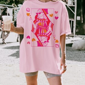 May include: A light pink t-shirt featuring a graphic of a queen playing card in shades of pink and orange. The word "BLOSSOM" is printed in black at the bottom of the shirt. The shirt has short sleeves and a relaxed fit.