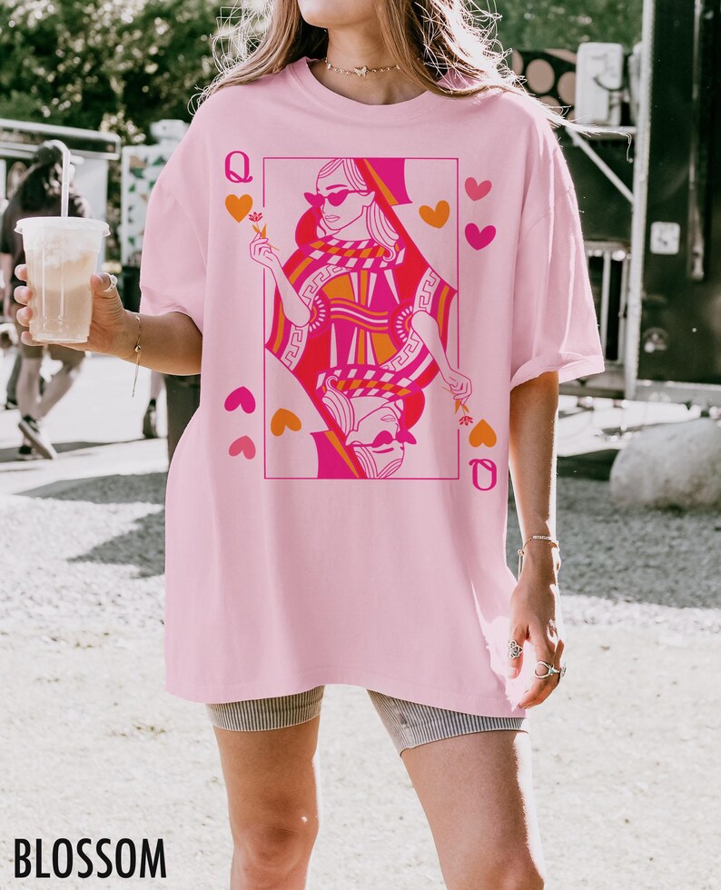 May include: A light pink t-shirt featuring a graphic of a queen playing card in shades of pink and orange. The word "BLOSSOM" is printed in black at the bottom of the shirt. The shirt has short sleeves and a relaxed fit.