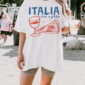 K&ouml;nnte beinhalten: Wei&szlig;es Oversize-T-Shirt mit einem Aufdruck eines Weinglases und einem St&uuml;ck Pizza. Der Text "ITALIA vino e pizza" ist auf das Shirt gedruckt.