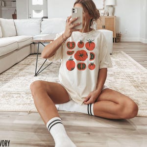 May include: A white t-shirt featuring a graphic of red tomatoes. The shirt has a relaxed fit and is shown being worn by a person with the text "IVORY" in the bottom left corner of the image.