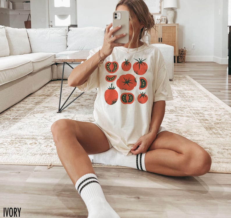 May include: A white t-shirt featuring a graphic of red tomatoes. The shirt has a relaxed fit and is shown being worn by a person with the text "IVORY" in the bottom left corner of the image.