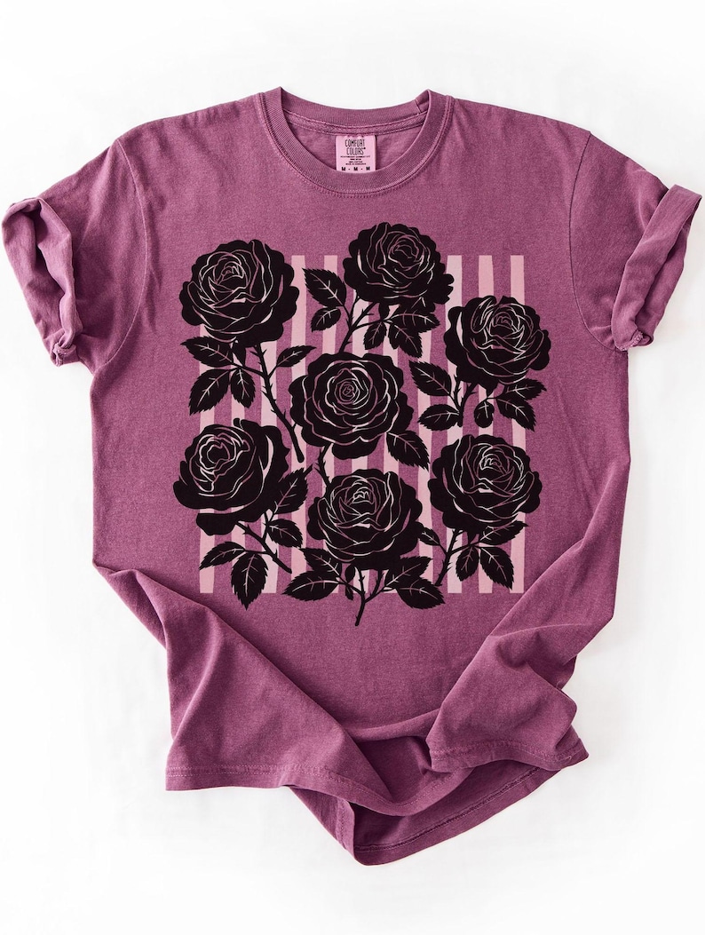 May include: A mauve t-shirt featuring a black rose graphic. The design includes seven black roses with stems and leaves, set against a background of vertical pink and white stripes. The shirt has short sleeves and a crew neck.
