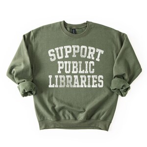 May include: An olive green sweatshirt with the text "SUPPORT PUBLIC LIBRARIES" in distressed white lettering. The crew neck sweatshirt has long sleeves with rolled cuffs. The image is on a white background.