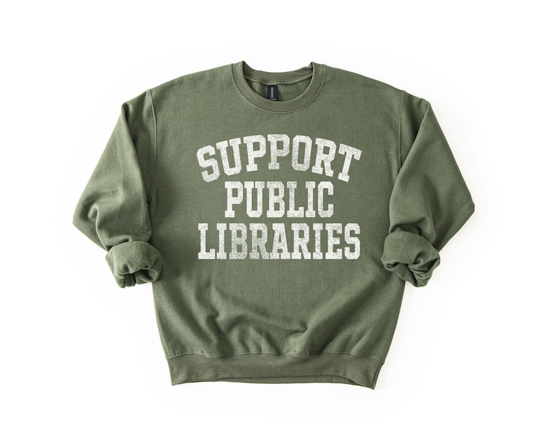 May include: An olive green sweatshirt with the text "SUPPORT PUBLIC LIBRARIES" in distressed white lettering. The crew neck sweatshirt has long sleeves with rolled cuffs. The image is on a white background.