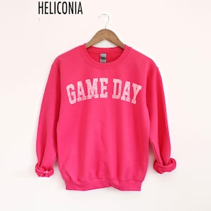 May include: A bright pink sweatshirt with the text "GAME DAY" in white block letters. The sweatshirt is hanging on a wooden hanger against a white background. The brand name "HELICONIA" is printed at the top.