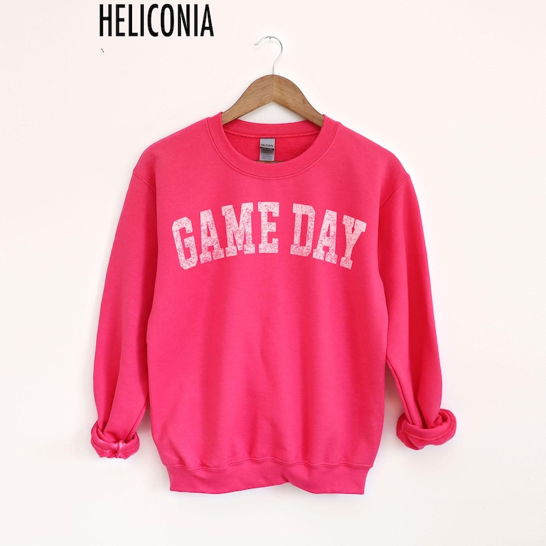 May include: A bright pink sweatshirt with the text "GAME DAY" in white block letters. The sweatshirt is hanging on a wooden hanger against a white background. The brand name "HELICONIA" is printed at the top.