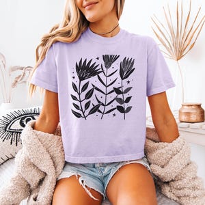 May include: A lilac t-shirt with a black floral print, including leaves and stars. The short-sleeved top has a round neck. The model is wearing denim shorts and a beige cardigan.