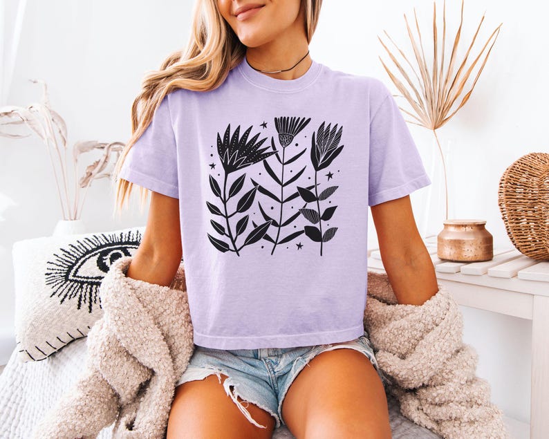 May include: A lilac t-shirt with a black floral print, including leaves and stars. The short-sleeved top has a round neck. The model is wearing denim shorts and a beige cardigan.