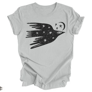 Peut inclure: Un t-shirt argent&eacute; avec un motif noir stylis&eacute; d'un oiseau avec des &eacute;toiles et une lune croissante. L'oiseau vole vers la droite.