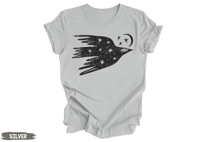 Peut inclure: Un t-shirt argent&eacute; avec un motif noir stylis&eacute; d'un oiseau avec des &eacute;toiles et une lune croissante. L'oiseau vole vers la droite.