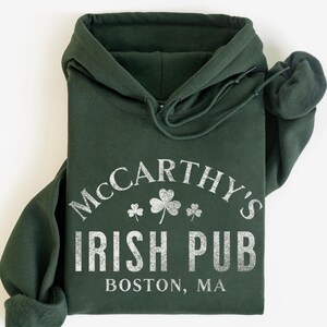 May include: Dark green hooded sweatshirt featuring the text "McCarthy's Irish Pub Boston, MA" in a distressed white font, with shamrock graphics. The sweatshirt has a drawstring hood and cuffed sleeves.