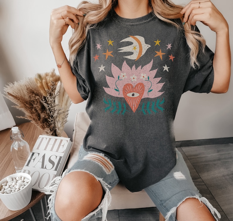 May include: Dark grey t-shirt with a celestial design. The graphic features a bird, crescent moon, stars, and a heart with an eye. The design uses pink, orange, yellow, and teal. The shirt has a relaxed fit.