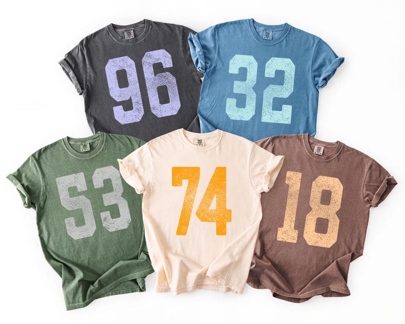 May include: Five vintage-style t-shirts in various colors, each displaying a large number. The shirts are in shades of charcoal, blue, green, cream, and brown, with numbers 96, 32, 53, 74, and 18 printed in contrasting colors. The shirts have rolled-up sleeves.