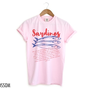 May include: Pink t-shirt with a blue and red illustration of sardines and the text "Sardines" in red. The text below the illustration reads "Oh, witness the  discerning palates of the discerning... a descent from the world waters of the Mediterranean... a diet... from the world's only source of being bold... where the waves build their sandy paths... Got it? You're a...  a simple pleasure and a handsomely presented...  a mouthful of goodness and culinary finesse... a reminder of...  a simple pleasure and a mighty mouthful... the messy...  sardines will go on forever in our hearts...  of the sea!"