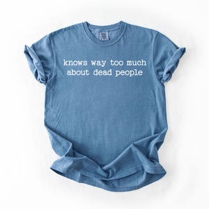 Puede incluir: Una camiseta azul con el texto blanco "knows way too much about dead people". La camiseta de manga corta es de color liso y se muestra plana sobre un fondo blanco.