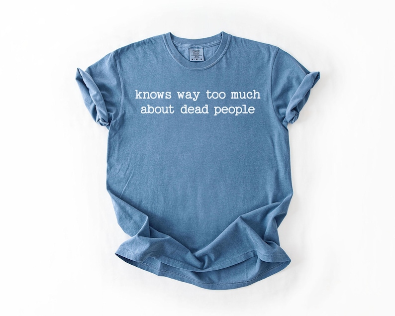 Puede incluir: Una camiseta azul con el texto blanco "knows way too much about dead people". La camiseta de manga corta es de color liso y se muestra plana sobre un fondo blanco.