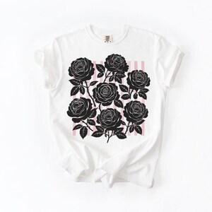 May include: White t-shirt featuring a black rose graphic design. The design includes seven black roses with stems and leaves, set against a backdrop of pink vertical stripes. The shirt has a classic crew neck and short sleeves.
