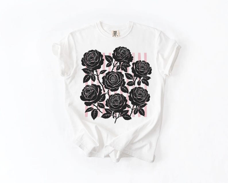 May include: White t-shirt featuring a black rose graphic design. The design includes seven black roses with stems and leaves, set against a backdrop of pink vertical stripes. The shirt has a classic crew neck and short sleeves.