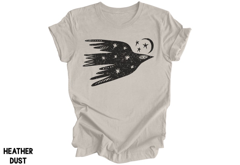 Peut inclure: Un t-shirt gris chin&eacute; avec un motif noir et blanc d'un oiseau stylis&eacute; volant avec des &eacute;toiles et une lune croissante.