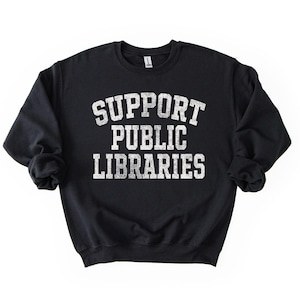 May include: Black crew neck sweatshirt with the text "SUPPORT PUBLIC LIBRARIES" in distressed white lettering. The sweatshirt is a solid black color and has long sleeves.