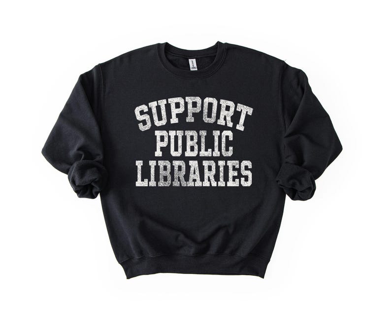 May include: Black crew neck sweatshirt with the text "SUPPORT PUBLIC LIBRARIES" in distressed white lettering. The sweatshirt is a solid black color and has long sleeves.