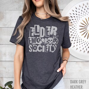 May include: A dark grey heather t-shirt with the text "Elder Emo Society" printed in white. The text is stylized to look like cut-out letters with a distressed effect. The design also includes a skull and crossbones graphic.