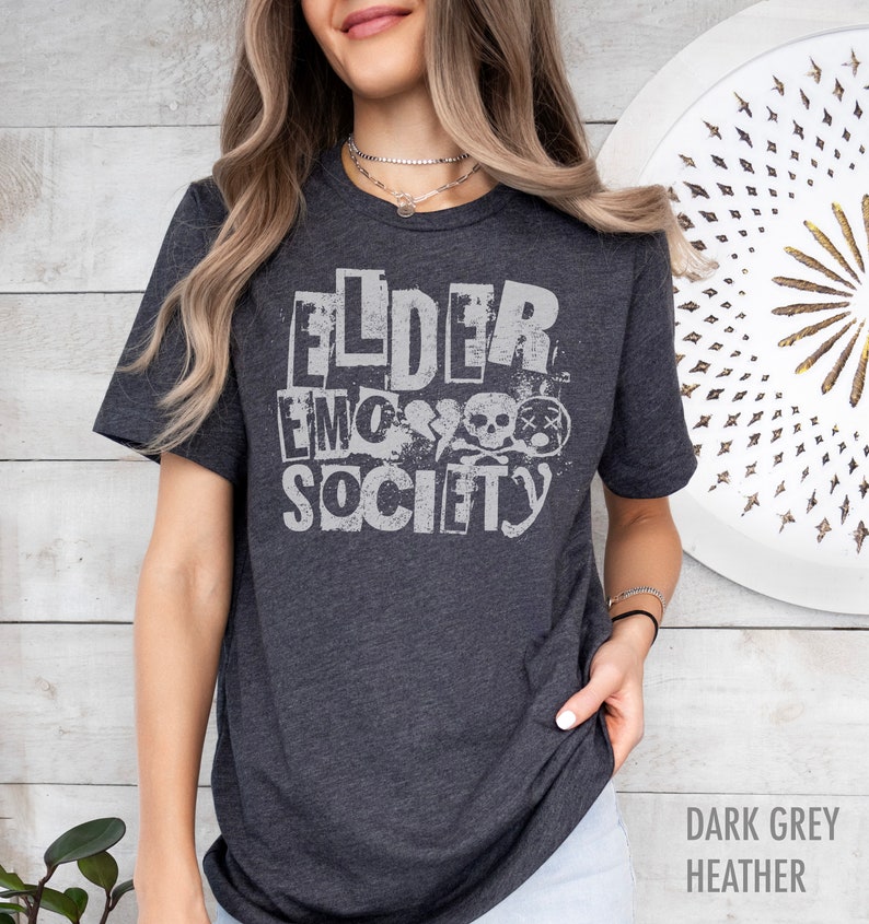 May include: A dark grey heather t-shirt with the text "Elder Emo Society" printed in white. The text is stylized to look like cut-out letters with a distressed effect. The design also includes a skull and crossbones graphic.