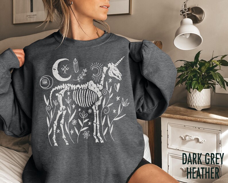 May include: Dark grey heather sweatshirt featuring a white graphic of a unicorn skeleton with celestial and floral elements. The design includes a crescent moon, sun, and various botanical details. The text "DARK GREY HEATHER" is visible.