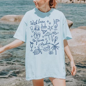 Puede incluir: Camiseta azul claro con un gr&aacute;fico de iconos de playa y el texto "With love from Aruba" en azul oscuro. El dise&ntilde;o incluye un cangrejo, un bikini, una estrella de mar y otras ilustraciones relacionadas con las vacaciones.