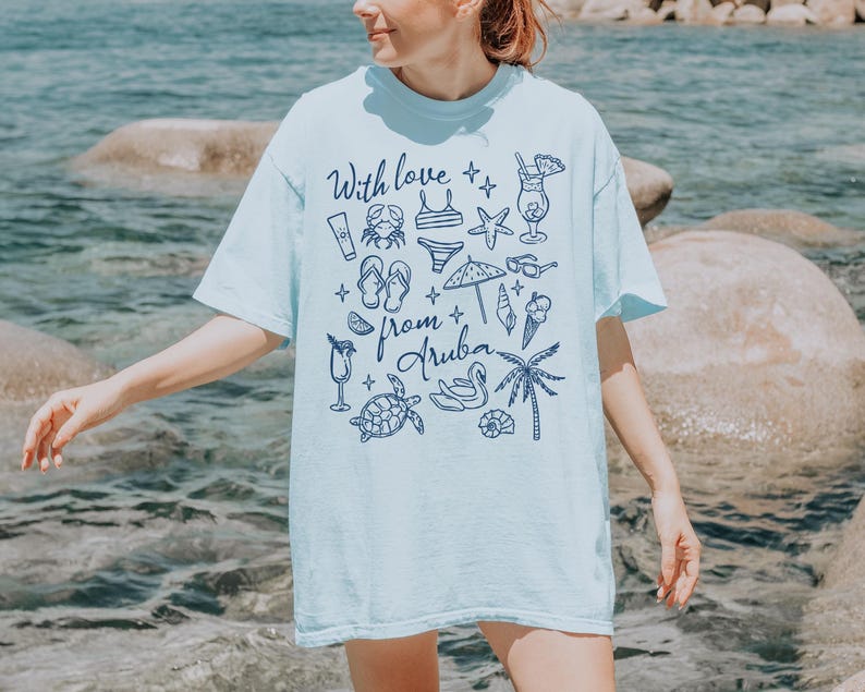 Puede incluir: Camiseta azul claro con un gr&aacute;fico de iconos de playa y el texto "With love from Aruba" en azul oscuro. El dise&ntilde;o incluye un cangrejo, un bikini, una estrella de mar y otras ilustraciones relacionadas con las vacaciones.