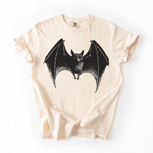 May include: A cream-colored t-shirt featuring a detailed black illustration of a bat with outstretched wings. The bat is centered on the shirt, showcasing its detailed features. The shirt has short sleeves and a crew neck.