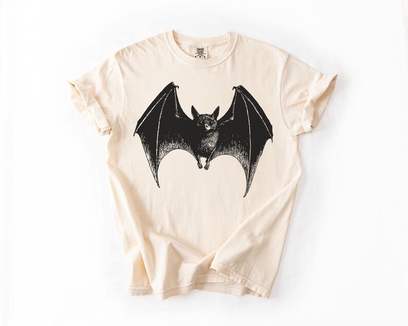 May include: A cream-colored t-shirt featuring a detailed black illustration of a bat with outstretched wings. The bat is centered on the shirt, showcasing its detailed features. The shirt has short sleeves and a crew neck.