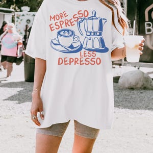 May include: White t-shirt with a blue and red graphic. The graphic features a coffee cup, a stovetop espresso maker, and the text "MORE ESPRESSO LESS DEPRESSO". The shirt is a casual, oversized style.