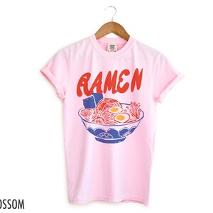 May include: Pink t-shirt featuring a graphic of a bowl of ramen noodles and the word "Ramen" in red lettering.