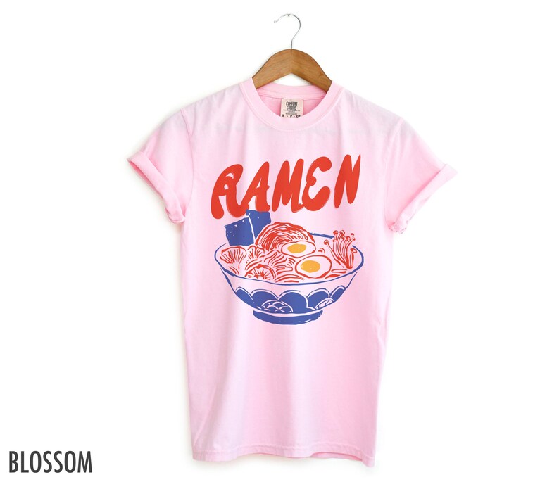 May include: Pink t-shirt featuring a graphic of a bowl of ramen noodles and the word "Ramen" in red lettering.