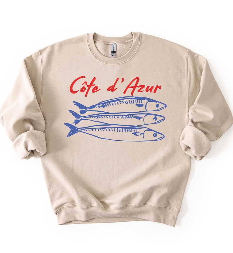 K&ouml;nnte beinhalten: Beiges Sweatshirt mit dem roten Schriftzug "C&ocirc;te d'Azur" &uuml;ber drei blauen Fischillustrationen. Das Sweatshirt hat einen Rundhalsausschnitt und lange &Auml;rmel.