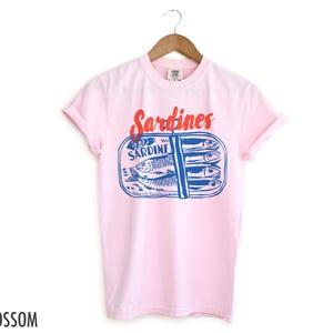 K&ouml;nnte beinhalten: Pinkes T-Shirt mit einem Aufdruck einer Sardinenb&uuml;chse. Die Dose tr&auml;gt die Aufschrift "WC Sardine" und "Genuine Pilchards in Oil". Der Text "Sardines" ist rot.