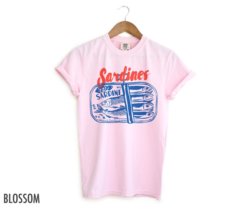 K&ouml;nnte beinhalten: Pinkes T-Shirt mit einem Aufdruck einer Sardinenb&uuml;chse. Die Dose tr&auml;gt die Aufschrift "WC Sardine" und "Genuine Pilchards in Oil". Der Text "Sardines" ist rot.