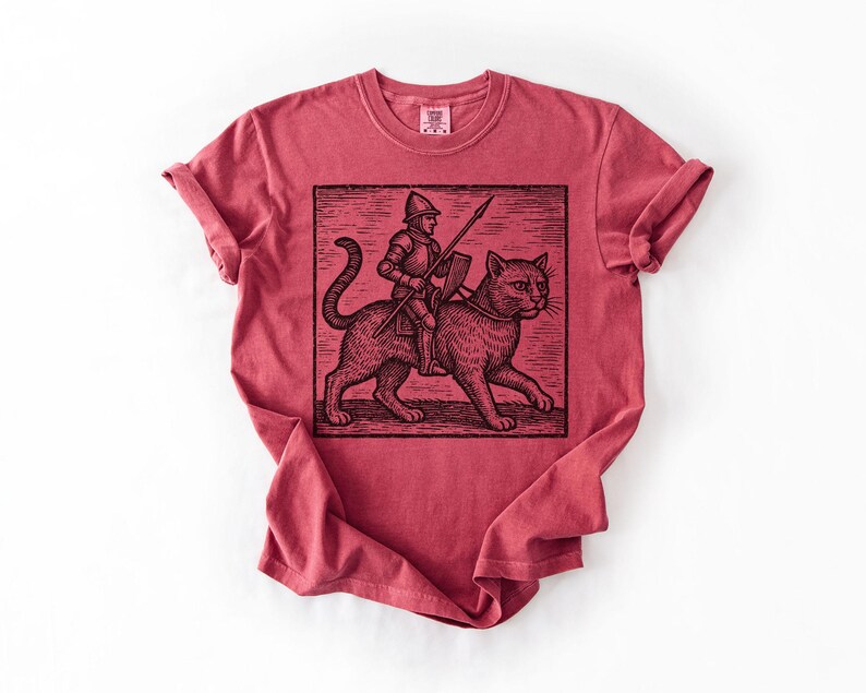 Puede incluir: Camiseta roja con una ilustraci&oacute;n en blanco y negro de un caballero montado en un gato, sosteniendo una lanza y un escudo. El gr&aacute;fico est&aacute; en un rect&aacute;ngulo. La camiseta tiene mangas enrolladas y cuello redondo.