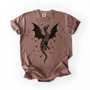 Op de afbeelding: Bruin t-shirt met een zwart drakenontwerp, uitgespreide vleugels en sterren. De draak staat centraal, met sterren eromheen. Het shirt heeft een ronde hals en korte mouwen. De stof lijkt zacht en comfortabel.
