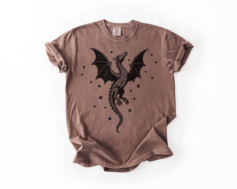 Op de afbeelding: Bruin t-shirt met een zwart drakenontwerp, uitgespreide vleugels en sterren. De draak staat centraal, met sterren eromheen. Het shirt heeft een ronde hals en korte mouwen. De stof lijkt zacht en comfortabel.
