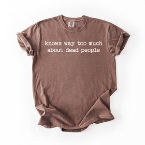 Puede incluir: Camiseta marr&oacute;n con el texto blanco "knows way too much about dead people". La camiseta tiene cuello redondo y mangas cortas. La tela tiene un aspecto ligeramente descolorido.