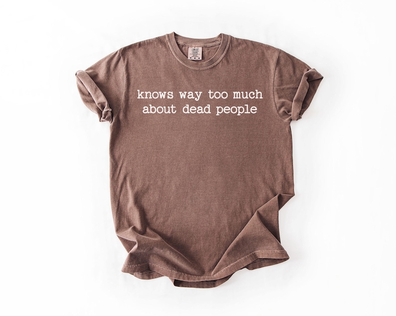 Puede incluir: Camiseta marr&oacute;n con el texto blanco "knows way too much about dead people". La camiseta tiene cuello redondo y mangas cortas. La tela tiene un aspecto ligeramente descolorido.