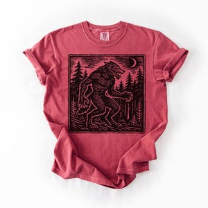 Puede incluir: Camiseta roja con un dise&ntilde;o negro de un hombre lobo en un bosque bajo una luna creciente. El dise&ntilde;o est&aacute; enmarcado dentro de un borde rectangular. La camiseta tiene mangas enrolladas y cuello redondo.