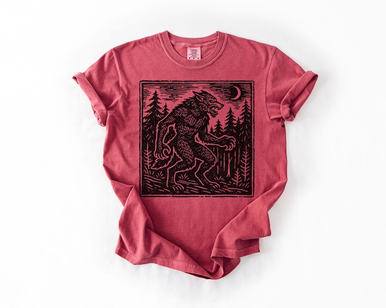 Puede incluir: Camiseta roja con un dise&ntilde;o negro de un hombre lobo en un bosque bajo una luna creciente. El dise&ntilde;o est&aacute; enmarcado dentro de un borde rectangular. La camiseta tiene mangas enrolladas y cuello redondo.