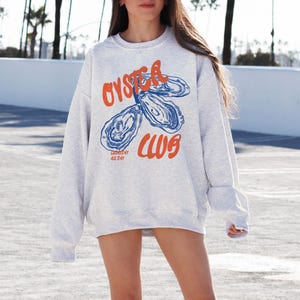 K&ouml;nnte beinhalten: Ein graues Sweatshirt mit einer blauen und orangenen Grafik von Austern und dem Text "Oyster Club" mit dem Slogan "Good Times All Day".