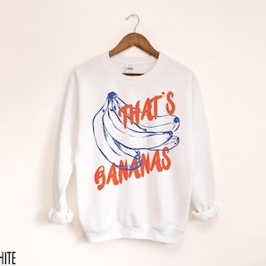 K&ouml;nnte beinhalten: Wei&szlig;er Pullover mit einer blauen und orangenen Grafik von Bananen und dem Text "That's Bananas".