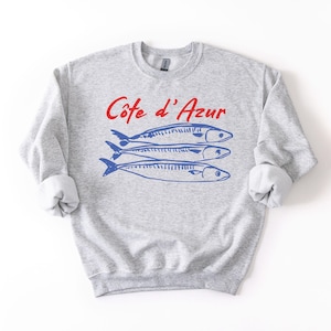 K&ouml;nnte beinhalten: Hellgraues Sweatshirt mit hochgekrempelten &Auml;rmeln. Auf der Vorderseite steht der Text "C&ocirc;te d'Azur" in Rot &uuml;ber drei blauen Fischillustrationen. Ein l&auml;ssiges Kleidungsst&uuml;ck f&uuml;r den Alltag.