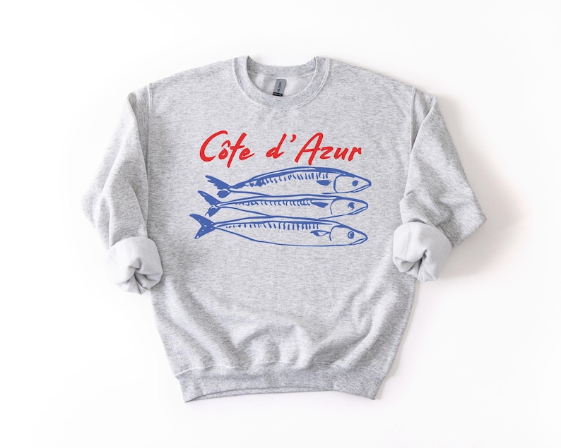 K&ouml;nnte beinhalten: Hellgraues Sweatshirt mit hochgekrempelten &Auml;rmeln. Auf der Vorderseite steht der Text "C&ocirc;te d'Azur" in Rot &uuml;ber drei blauen Fischillustrationen. Ein l&auml;ssiges Kleidungsst&uuml;ck f&uuml;r den Alltag.